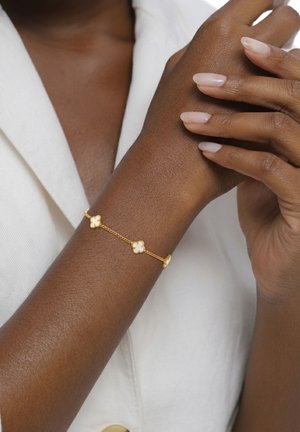 Mørk hudfarvet arm med et guldarmbånd med hvide kløverformede charms, hænder der blidt holder hinanden, hvidt tøj i baggrunden.