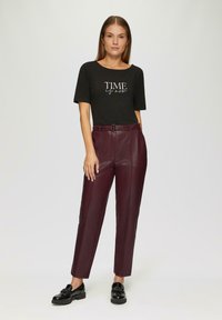 Zwarte T-shirt met witte "TIME" grafiek, gecombineerd met bordeaux kleurige neplederen broek. Model draagt zwarte instappers. Eenvoudige achtergrond.