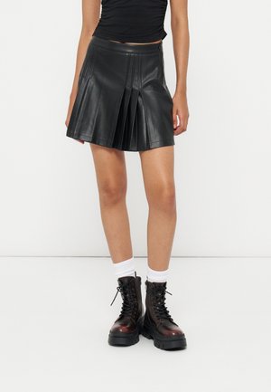Jeune personne portant une jupe en cuir noire plissée, des chaussettes blanches et des bottes de combat marron foncé à lacets, debout devant un fond blanc uni.