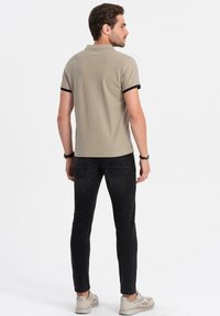 Béžové polo tričko s černým límcem a rukávy, spárované s černými džíny. Model stojí zády, předvádějící štíhlý střih.