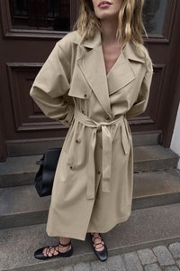 Bežs tranšejas mētelis ar dubultajiem pogām, plašām piedurknēm un jostu viduklī. Melni platais apavi ar krustveida siksnām. Redzama melna rokassoma.