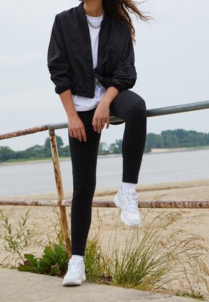 Veste bomber noire par-dessus un t-shirt blanc, associée à des leggings noirs avec des accents côtelés et des baskets blanches. Debout sur une rambarde.