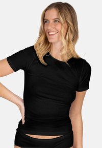 Smiling woman with blonde hair wearing a fitted black T-shirt and black underwear, posing with hand on hip against white background.
