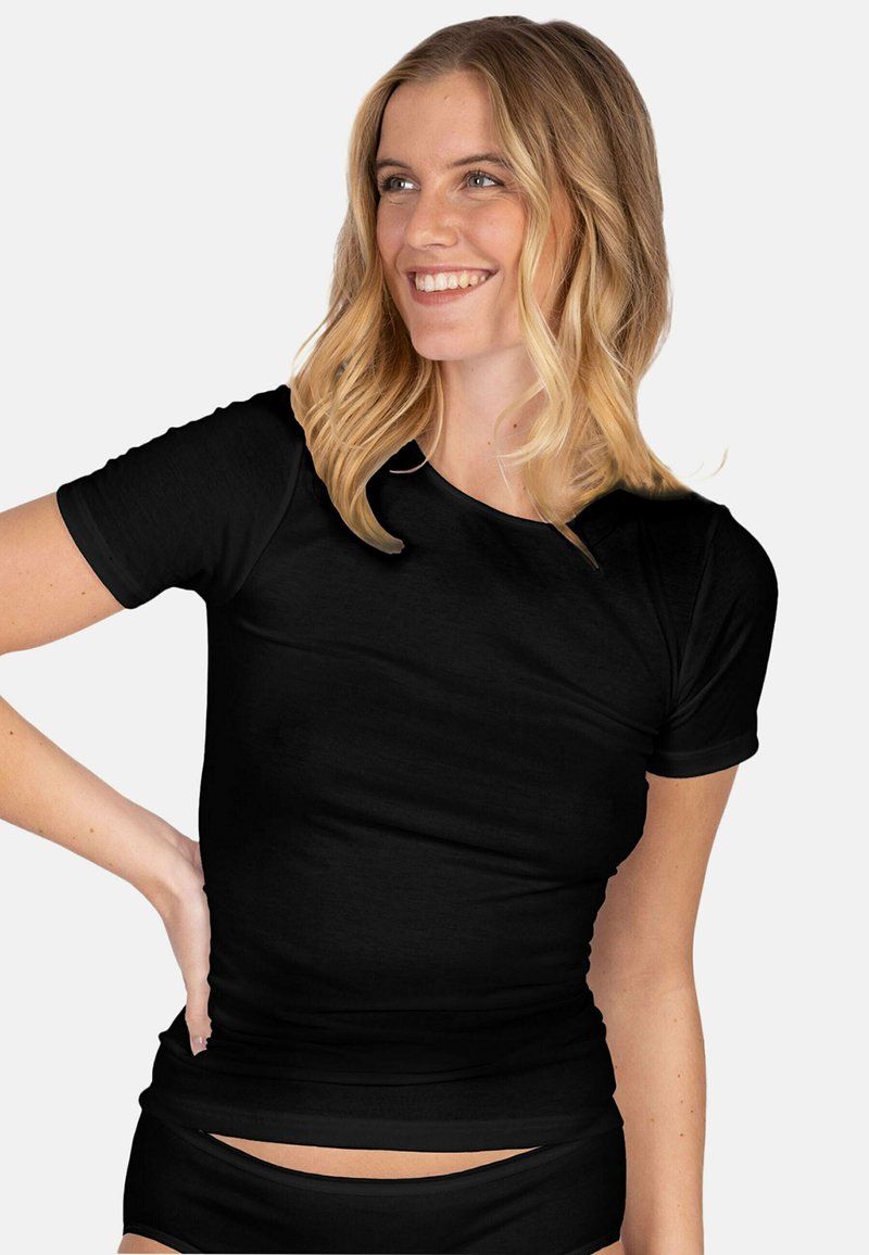Smiling woman with blonde hair wearing a fitted black T-shirt and black underwear, posing with hand on hip against white background.