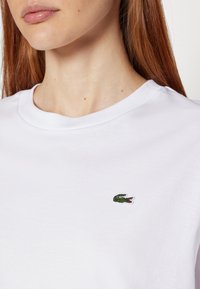 Weißes Baumwoll-T-Shirt mit Rundhalsausschnitt, versehen mit einem kleinen grünen Krokodil-Logo, das auf der Brust gestickt ist. Glatte Textur.