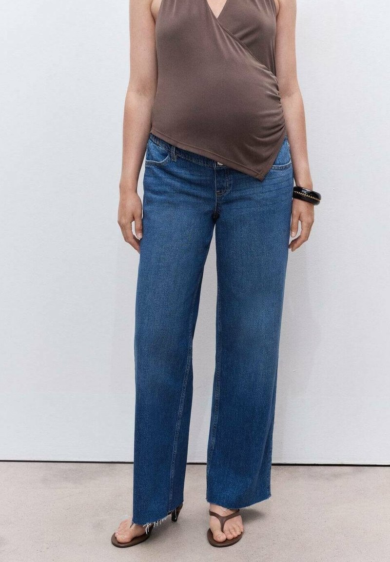 Personne portant un top cache-cœur marron sans manches, un jean bleu à jambes larges avec des ourlets effilochés, un bracelet noir et des sandales marron à talons, debout contre un mur blanc.