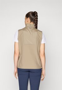Beige, sleeveless vest with a high collar and a smooth texture, designed for a fitted look; worn over a white T-shirt, paired with navy pants.