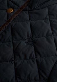 Gros plan sur un tissu matelassé marine avec des bords en cordon marron, un bouton-pression en laiton et un subtil détail de monogramme brodé.