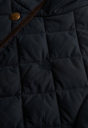 Gros plan sur un tissu matelassé marine avec des bords en cordon marron, un bouton-pression en laiton et un subtil détail de monogramme brodé.
