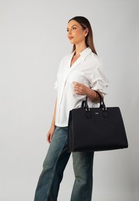 Black leather handbag with a structured design, featuring dual top handles and gold-tone hardware, held by a person in a white shirt and jeans.
