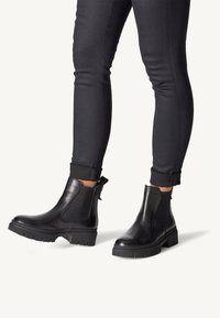 Botas Chelsea de cuero negro con suela gruesa, paneles laterales elásticos y tirador trasero, combinadas con pantalones negros ajustados.