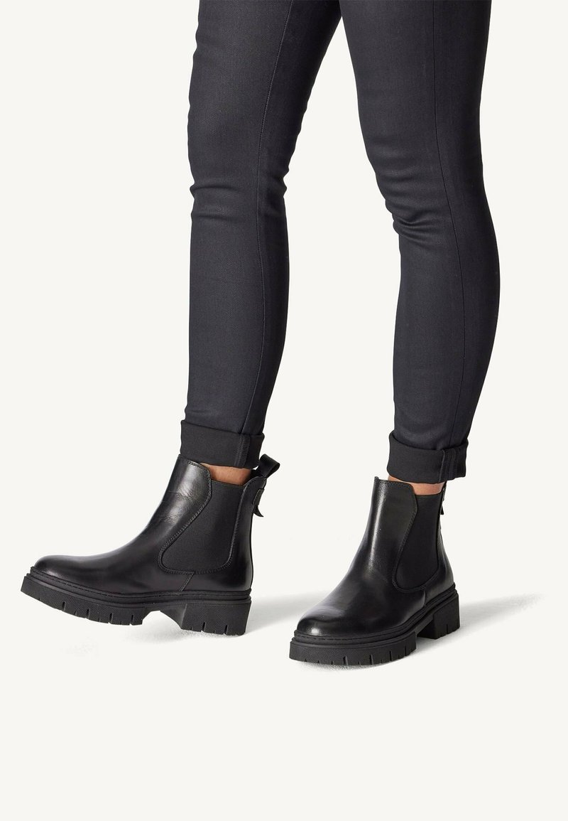 Botas Chelsea de cuero negro con suela gruesa, paneles laterales elásticos y tirador trasero, combinadas con pantalones negros ajustados.