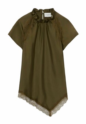 Femme portant une robe vert olive et des baskets noires tenant un sac à main bordeaux sur un fond blanc uni.