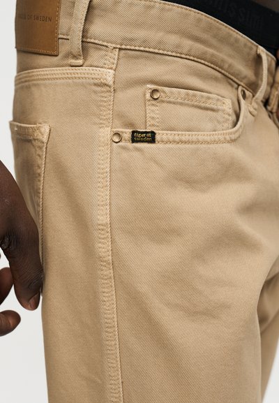 Beežid teksapüksid nähtavate õmblustega, väike esikülje münditasku ja „tiger of sweden“ silt tasku lähedal; tumedanahaline käsi kõrval.