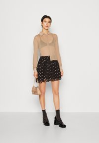 Camisa bege transparente com botões e mangas longas, combinada com uma saia preta com bolinhas douradas. Modelo segura uma pequena bolsa bege.