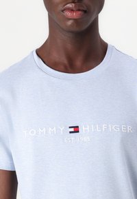 Světle modré bavlněné tričko s vyšitým logem "TOMMY HILFIGER" a designem vlajky, pod nímž je nápis "EST. 1985". Krátké rukávy, kulatý výstřih.