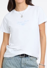 Camiseta de algodón blanca con cuello redondo, que presenta el logo en azul claro de "Guess Jeans" impreso en el frente. Combinada con shorts de mezclilla de cintura alta.