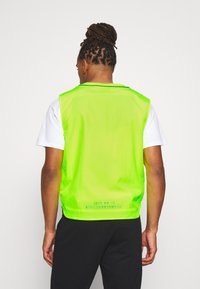 Neongrön ärmlös väst med en lätt och slät yta. Baksidan har text med varumärke i blått. Bärs över en vit skjorta.