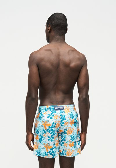 Άνδρας που στέκεται με την πλάτη στην κάμερα, φοράει πολύχρωμα μπλε και πορτοκαλί μαγιό με λουλούδια, με μια πίσω τσέπη και ετικέτα "Vilebrequin".