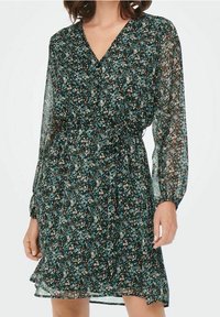 Donna che indossa un vestito a maniche lunghe, con scollatura a V, caratterizzato da un motivo floreale turchese, nero e bianco, e una vita aderente con una cintura di stoffa.