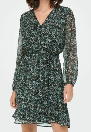 Frau in einem langärmligen, V-Ausschnitt-Kleid mit einem taubenblauen, schwarzen und weißen floralen Muster sowie einer taillierten Taille mit einem Stoffgürtel.