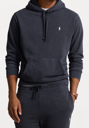 Mann trägt dunkelblauen Hoodie und Jogginghose mit kleinem weißem Polo-Logo, Hände entspannt, eine Hand in der Tasche, weißes Unterhemd am Hals sichtbar.
