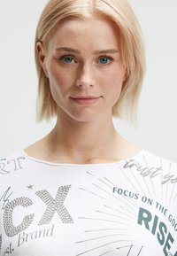 Blonde Frau mit blauen Augen und Sommersprossen, die ein weißes Oberteil mit grafischem Text und Sternenburst-Design trägt, frontal vor einem schlichten Hintergrund steht.