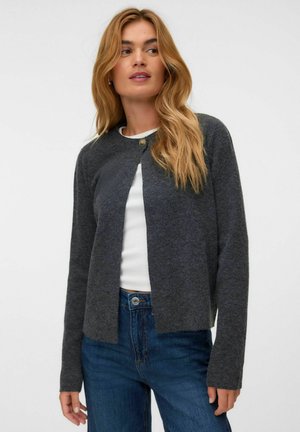 Donna con lunghi capelli biondi che indossa un cardigan grigio scuro abbottonato sopra una maglietta bianca e jeans blu, in piedi davanti a uno sfondo chiaro uniforme.