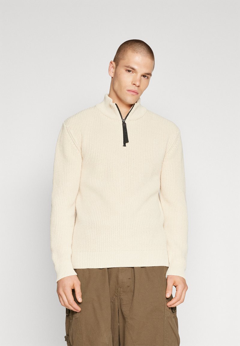Suéter de punto en color crema con cuello de media cremallera y patrón texturizado; combinado con pantalones cargo marrones.