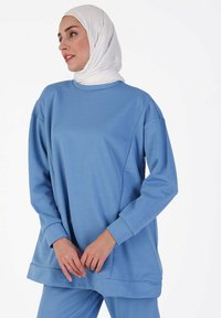 Light blue long-sleeve tunic made of soft fabric, featuring a crew neckline, dropped shoulders, and a relaxed fit. Smooth texture with no patterns.
