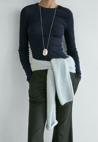 Marineblaues Langarmoberteil, hellblauer Strickwickel am Taillenbereich und dunkel gestreifte Hose. Silberne Anhängerkette fügt einen metallischen Akzent hinzu.