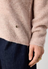 Gros plan d'une main avec une bague en or à côté d'un pull beige duveteux et d'un pantalon en denim bleu foncé.