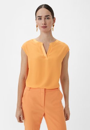 Orangenes, kurzärmeliges Blusenshirt mit V-Ausschnitt und glatter Textur, kombiniert mit passenden orangefarbenen maßgeschneiderten Hosen. Goldene Statement-Ohrringe.