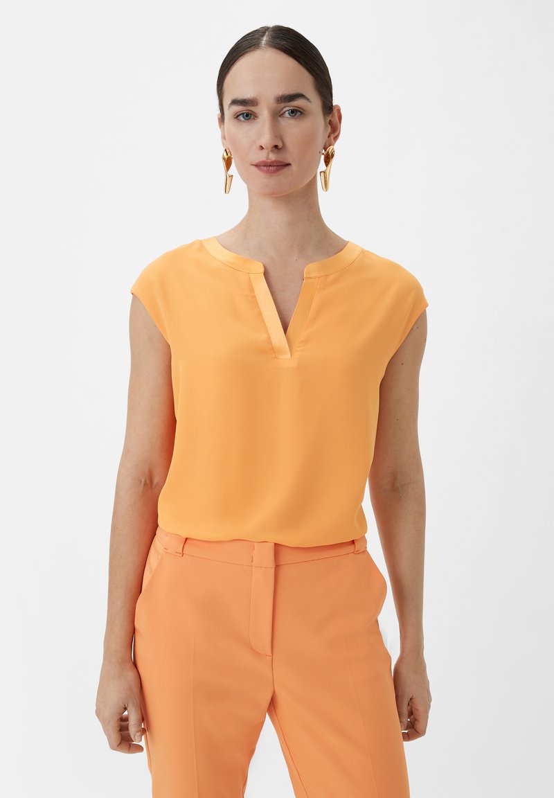 Orangenes, kurzärmeliges Blusenshirt mit V-Ausschnitt und glatter Textur, kombiniert mit passenden orangefarbenen maßgeschneiderten Hosen. Goldene Statement-Ohrringe.