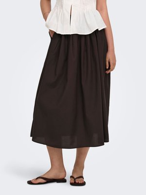 Femme portant une longue jupe marron foncé, un haut blanc sans manches et des sandales plates noires avec des lanières croisées, debout sur un fond uni.