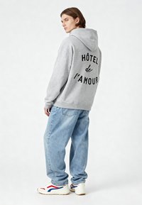 Szürke kapucnis pulóver, amelynek hátulján fekete szöveg olvasható: "HÔTEL de l'AMOUR". Bő kék farmernadrággal és piros és kék akcentusokkal ellátott fehér sportcipővel párosítva.