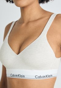 Απαλή εκρού μπραλέτα με V-λαιμόκοψη; μαλακή υφή, φαρδιές τιράντες και λευκή ελαστική ταινία με λογότυπο "Calvin Klein" σε μαύρο.