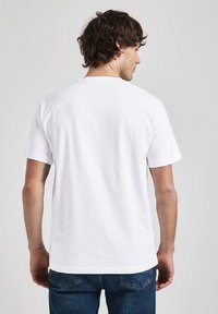 Bílý bavlněný tričko s krátkým rukávem, kulatým výstřihem a hladkým povrchem, zobrazené zezadu, nošené s modrými džínami.