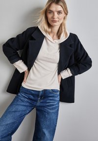 Marineblazer über einer cremefarbenen Kapuzenjacke mit gesticktem Text, kombiniert mit blauen Jeans. Glattes Material, lässige Passform, sichtbare Nähte.