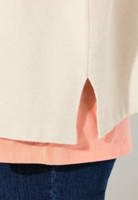 Miękki sweter w kolorze kremowym o teksturowanym wykończeniu, z asymetrycznym dołem i bocznymi rozcięciami, nałożony na jasnoróżową koszulkę i ciemne dżinsy.