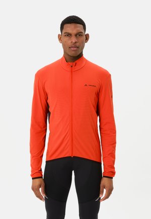 Mann trägt eine leuchtend orangefarbene Sportjacke mit Reißverschluss und schwarze Fahrradbekleidung, steht vor einem schlichten weißen Hintergrund.