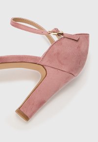 Chaussure à talon aiguille en suede rose avec une bride de cheville, bout pointu et talon épais. Présente un petit accent en boucle dorée.