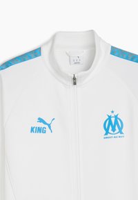 Vit zip-up träningsjacka med blå detaljer, logotyp och texten "KING". Inkluderar en geometrisk mönstrad kant vid axlarna.
