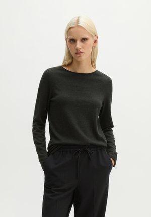 Femme blonde portant un pull à manches longues foncé et un pantalon noir à cordon, debout les mains dans les poches devant un fond uni.