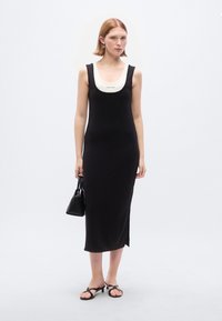 Μαύρο αμάνικο φόρεμα με λευκή λαιμόκοψη σε σχήμα στρογγυλής λεκάνης, πλευρική σχισμή, κατασκευασμένο από λείο υλικό. Το μοντέλο κρατά μια μικρή μαύρη τσάντα και φορά επίπεδα σανδάλια.