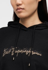 Μαύρο φούτερ με κουκούλα και ανάγλυφο σχέδιο από χρυσές στρας. Διαθέτει κουκούλα με κορδόνι και μεταλλικές λεπτομέρειες στα άκρα του κορδονιού.