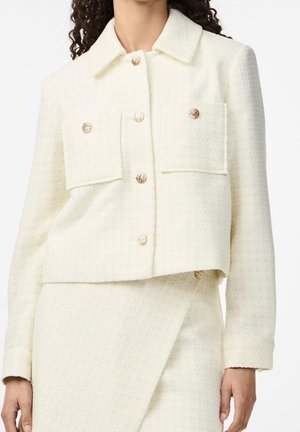 Femme portant une veste écrue texturée avec des boutons dorés et une jupe assortie avec un panneau avant superposé.