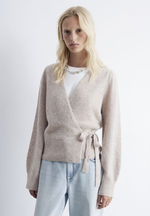 Light beige wrap sweater in soft, fluffy material with long sleeves and a tie closure; worn over a white shirt and paired with blue jeans.