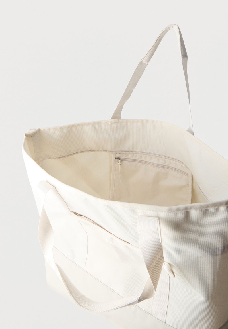 Große cremefarbene Tragetasche mit langen Griffen und innenliegender Reißverschlusstasche, offen gezeigt vor einfarbigem Hintergrund.