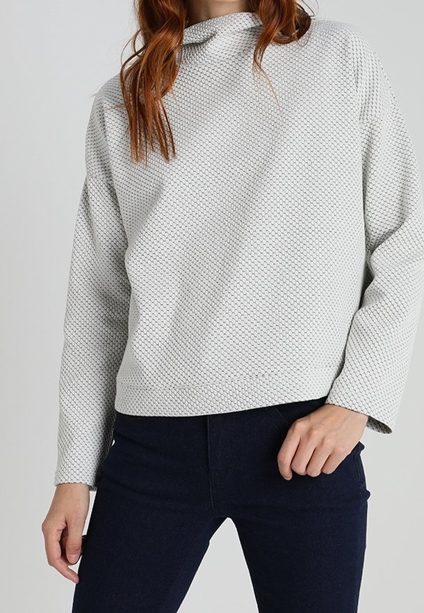 Pull gris clair texturé avec une coupe carrée, manches longues et col montant, associé à un jean foncé ajusté.